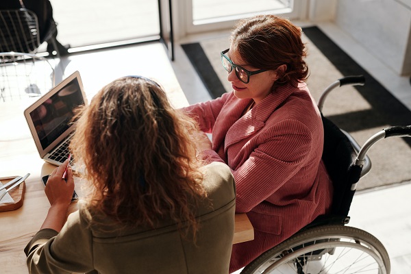 two women sitting at a laptop talking, one in a wheelchair and one sitting in a chair